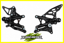 A pair of black metal motorcycle foot pegs with silver bolts, displayed on a white background with a yellow border and a 'UK ROAD AND RACE' logo at the bottom.